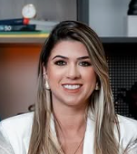 Mulher de pele clara, cabelos longos em tons de loiro e castanho, sorrindo para a câmera. Ela usa maquiagem social leve, brincos dourados e um blazer branco. O fundo é um ambiente de escritório moderno levemente desfocado.
